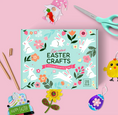 Load image into Gallery viewer, A flat-lay of an Easter craft kit box on a pink background, surrounded by themed props including scissors, wooden sticks, a gnome, a chick cutout, and painted Easter eggs. The box reads 'Easter Crafts' and shows white rabbits, flowers, and leaves on a light blue background.