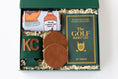 Load image into Gallery viewer, A curated golf gift set in a dark green box filled with shredded paper, featuring a rolled white kitchen towel with whiskey bottle graphics, a green drink sweater with 'KC' in gold, two brown leather coasters, and a green book titled 'The Golf Bucket List' by Jeff Thoreson.