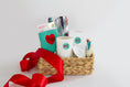 Load image into Gallery viewer, A woven gift basket filled with assorted items including a white ceramic candle with a teal KC logo, a teal drink sweater with a red heart and KC Current themed Kitchen Towel and a red ribbon bow, photographed on a clean white background.