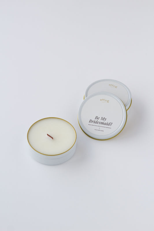 A white wax candle in a round white tin with a thin gold rim, shown next to one closed matching tin on a clean white background. One open tin label reads 'Be My Bridesmaid' and the brand 'effing candle co' is visible at the top.