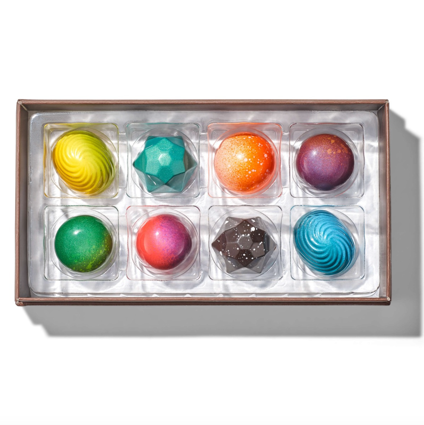 A boxed assortment of colorful chocolates in a clear tray with a metallic brown rim, photographed from above on a white background with a soft shadow.