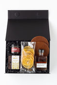 Load image into Gallery viewer, A curated gift box set in a black presentation box with shredded black filler, featuring a jar of cocktail cherries, a bag of California Crisps dried orange slices, a bottle of Strongwater Black Walnut cocktail bitters, a package of Andre's Bourbon Balls, and two brown leather coasters.
