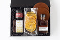 Load image into Gallery viewer, A curated gift box set in a black presentation box with shredded black filler, featuring a jar of cocktail cherries, a bag of California Crisps dried orange slices, a bottle of Strongwater Black Walnut cocktail bitters, a package of Andre's Bourbon Balls, and two brown leather coasters.
