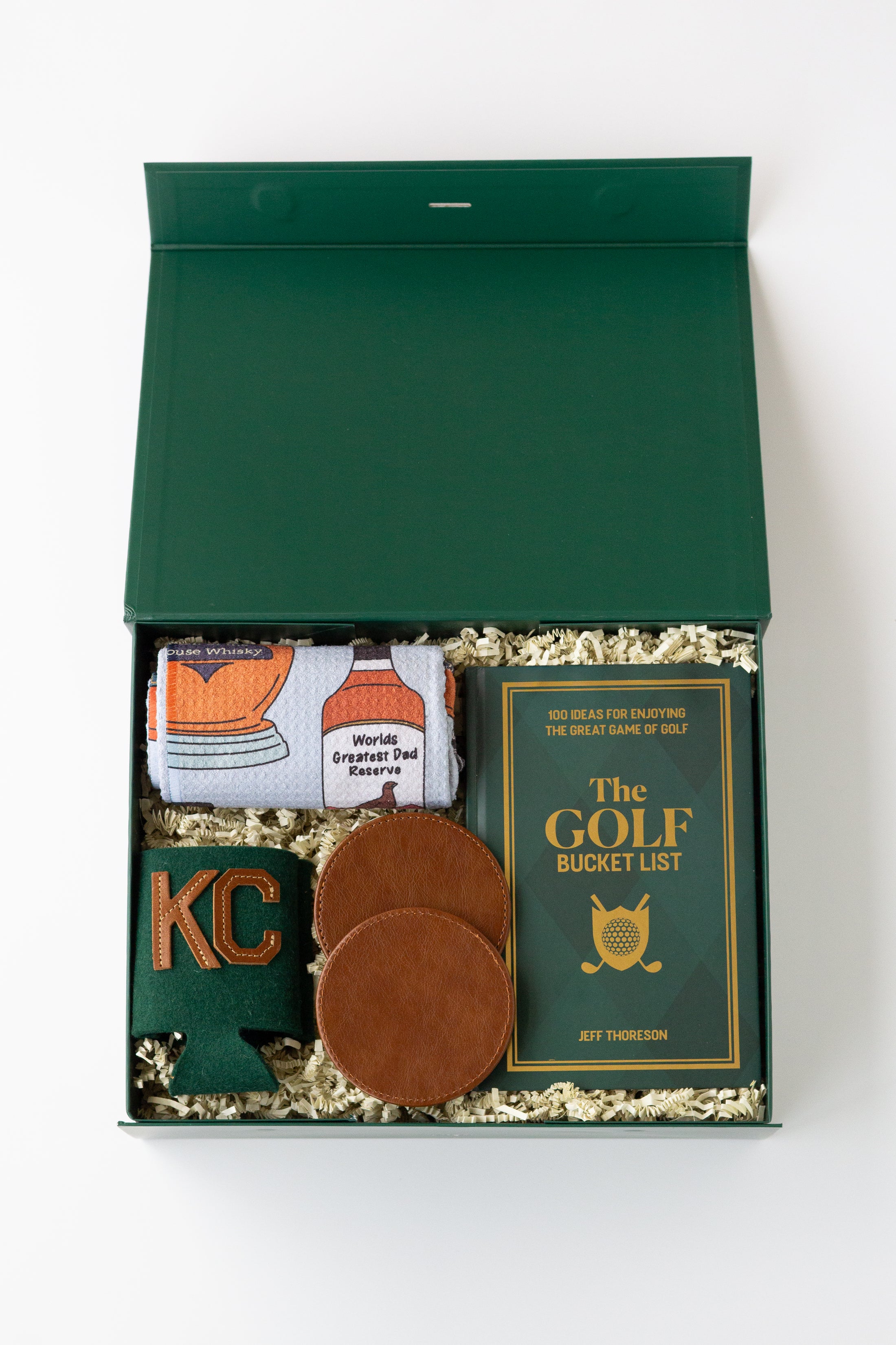 A curated golf gift set in a dark green box filled with shredded paper, featuring a rolled white kitchen towel with whiskey bottle graphics, a green drink sweater with 'KC' in gold, two brown leather coasters, and a green book titled 'The Golf Bucket List' by Jeff Thoreson.