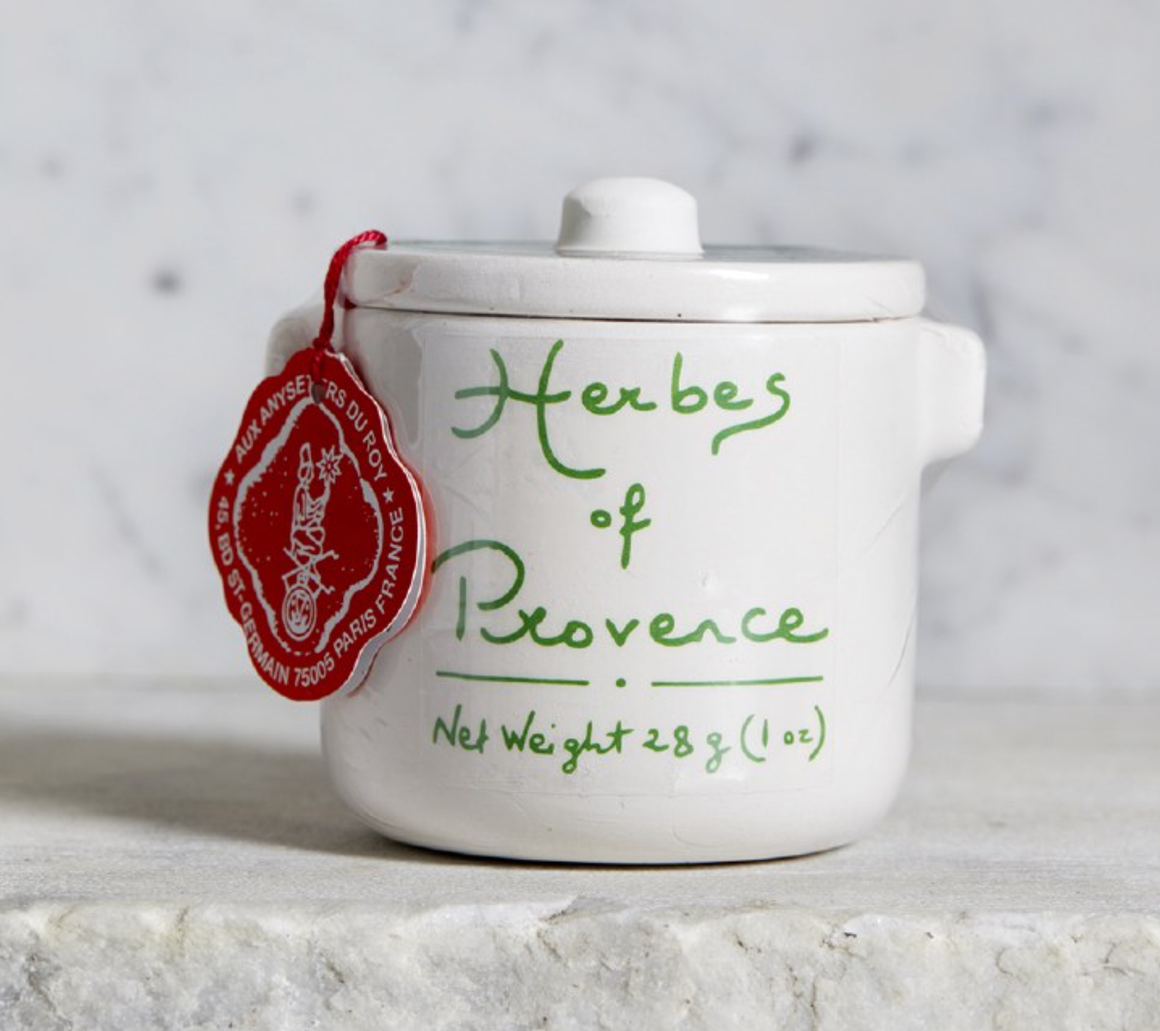 A small white ceramic jar with a lid, decorated with green cursive text reading 'Herbs of Provence' and a red hanging tag, set on a light stone surface against a soft marble-like background.