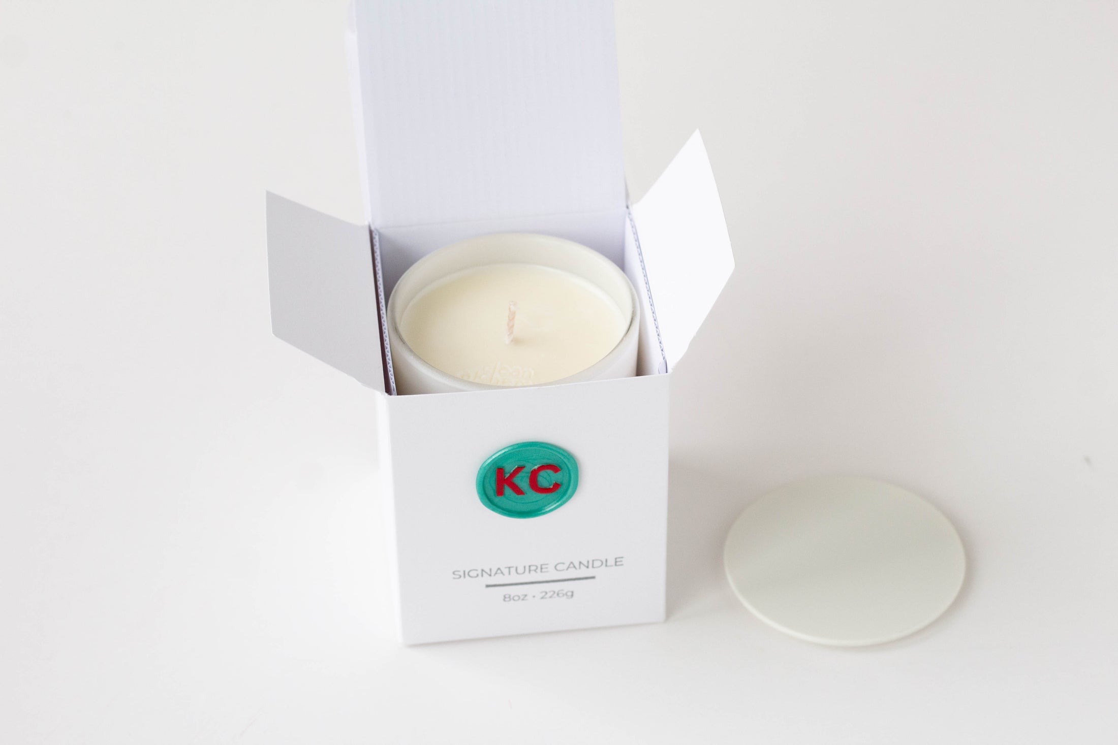 A white jar candle in an open white box with the lid placed beside it on a clean white background. The box has a green circular logo with red letters and the text 'KC' and 'SIGNATURE CANDLE'.