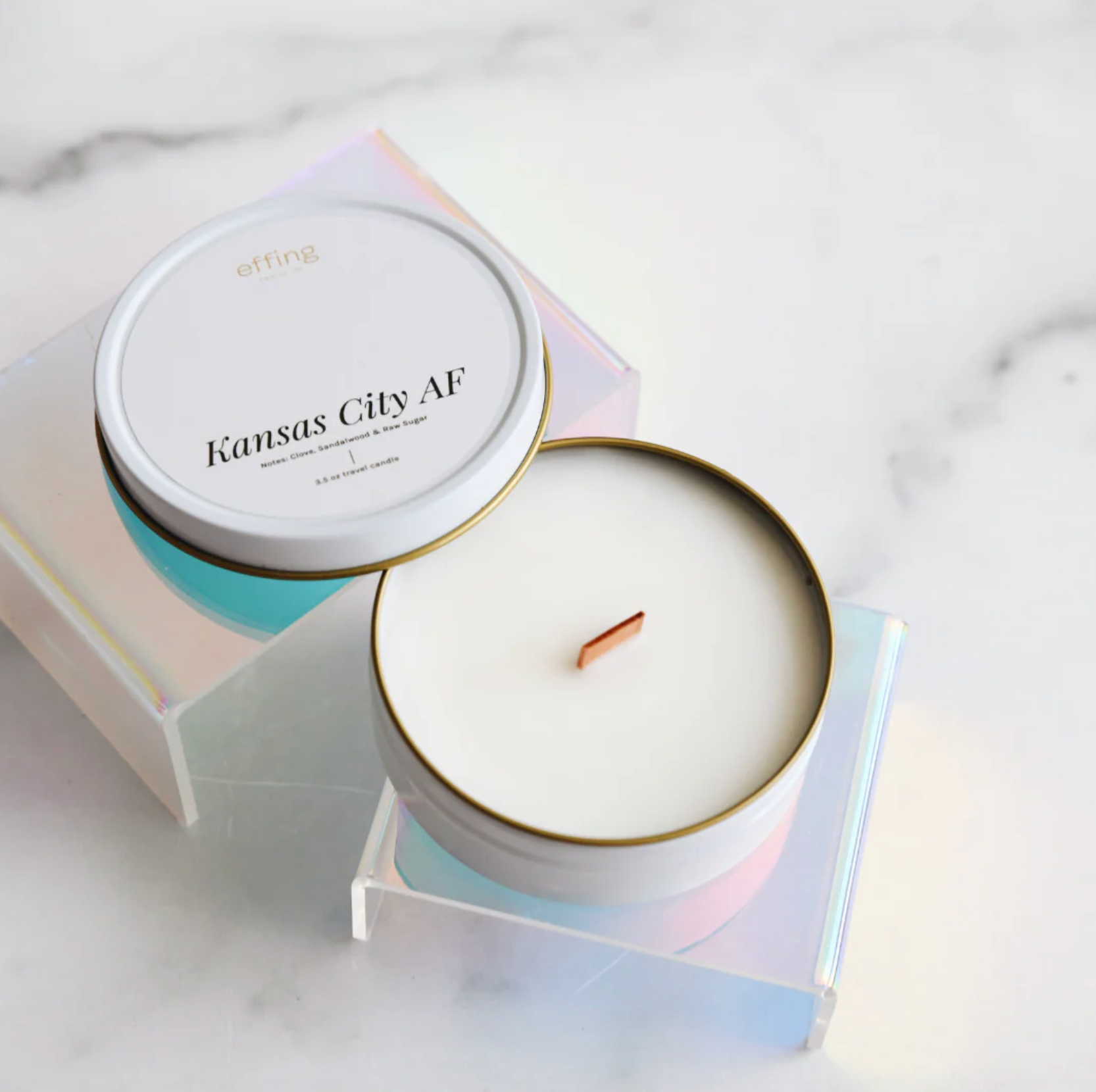 A white candle in a white travel tin sits on a reflective stand on a white marble surface. The candle label reads "Kansas City AF"