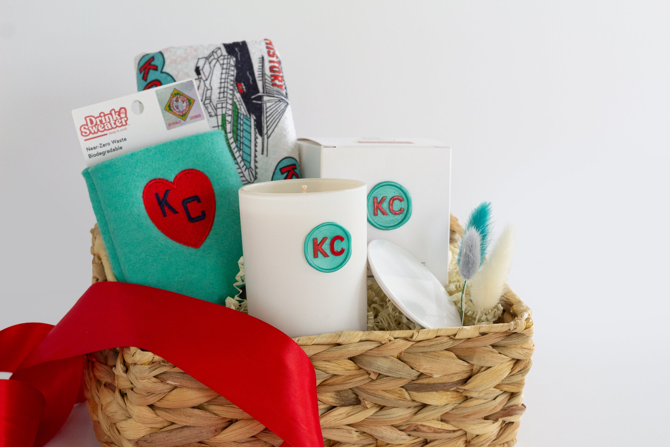 A woven gift basket filled with assorted items including a white ceramic candle with a teal KC logo, a teal drink sweater with a red heart and KC Current themed Kitchen Towel and a red ribbon bow, photographed on a clean white background.