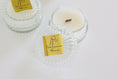 Load image into Gallery viewer, Two clear glass jar candles with gold 'Welcome Home' labels are arranged on a white surface. One candle is open, showing white wax and a small brown wood wick, while the other one is closed.
