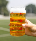 Load image into Gallery viewer, A hand holding a clear glass can-shaped mug filled with golden beer and topped with white foam. The mug has red 'KC' logos printed on it, and the background is a blurred outdoor field.
