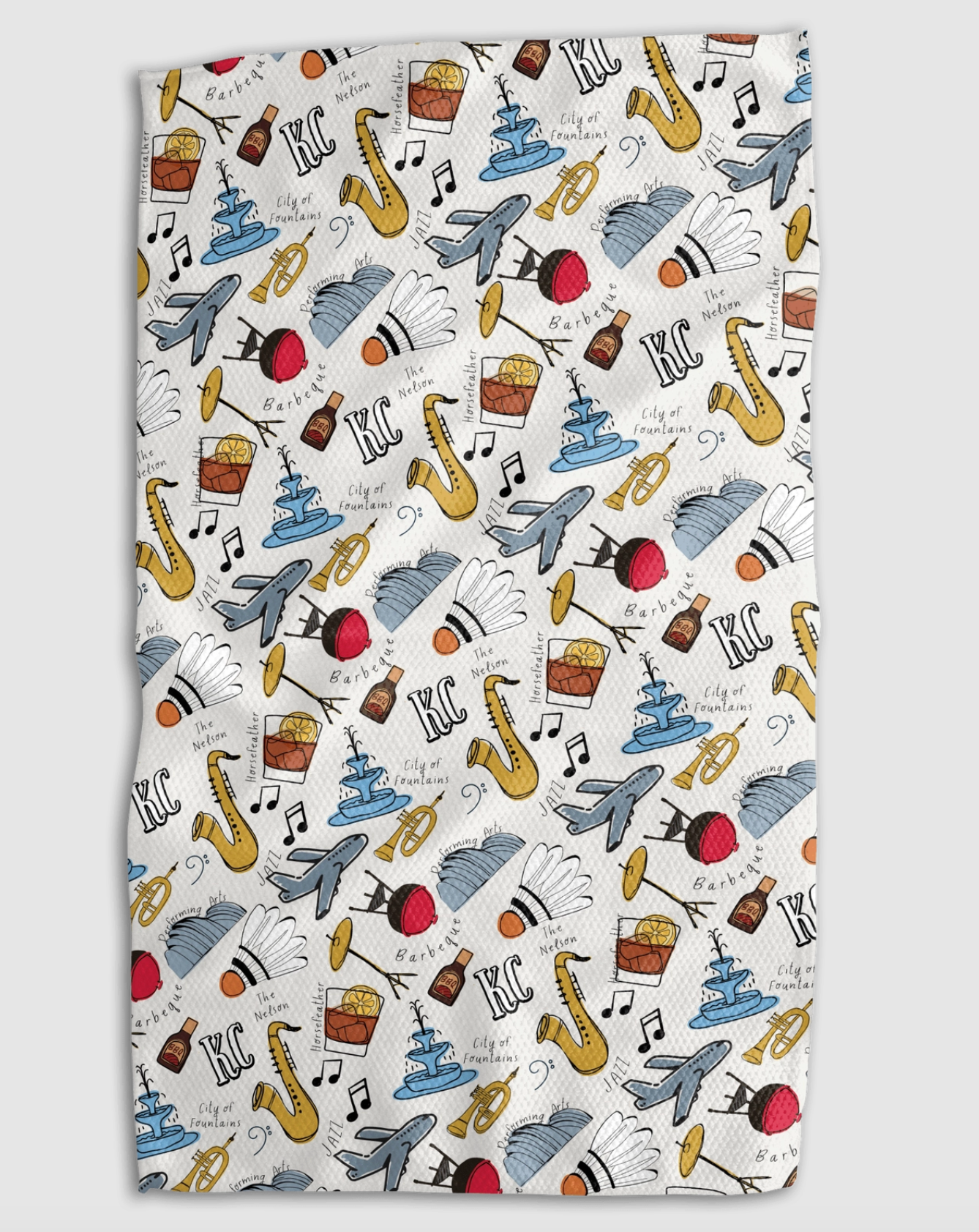 A rectangular kitchen towel with a white background and a colorful repeating pattern of Kansas City monuments, cocktail glasses, saxophones, BBQ sauce bottles, and the text 'KC'.