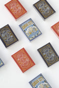 Load image into Gallery viewer, Several decks of playing cards arranged on a white background, showing different colorways and illustrated backs. 
