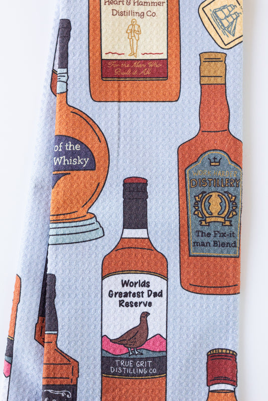 A light blue necktie featuring a repeating pattern of illustrated whiskey and spirit bottles in orange, brown, black, and cream, with humorous labels like 'World's Greatest Dad Reserve' and 'True Grit Distilling Co.'.