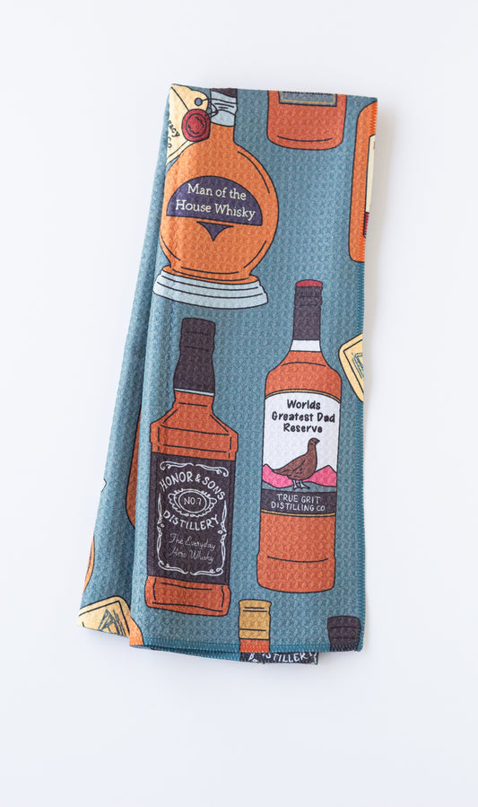 A folded towel, featuring a blue-green background with a pattern of illustrated whiskey bottles and labels in orange, black, white, and yellow.
