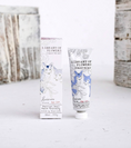 Load image into Gallery viewer, A tube and matching box of Library of Flowers Forget Me Not hand cream, featuring white packaging with blue and black botanical illustrations, set against a rustic white wooden background.

