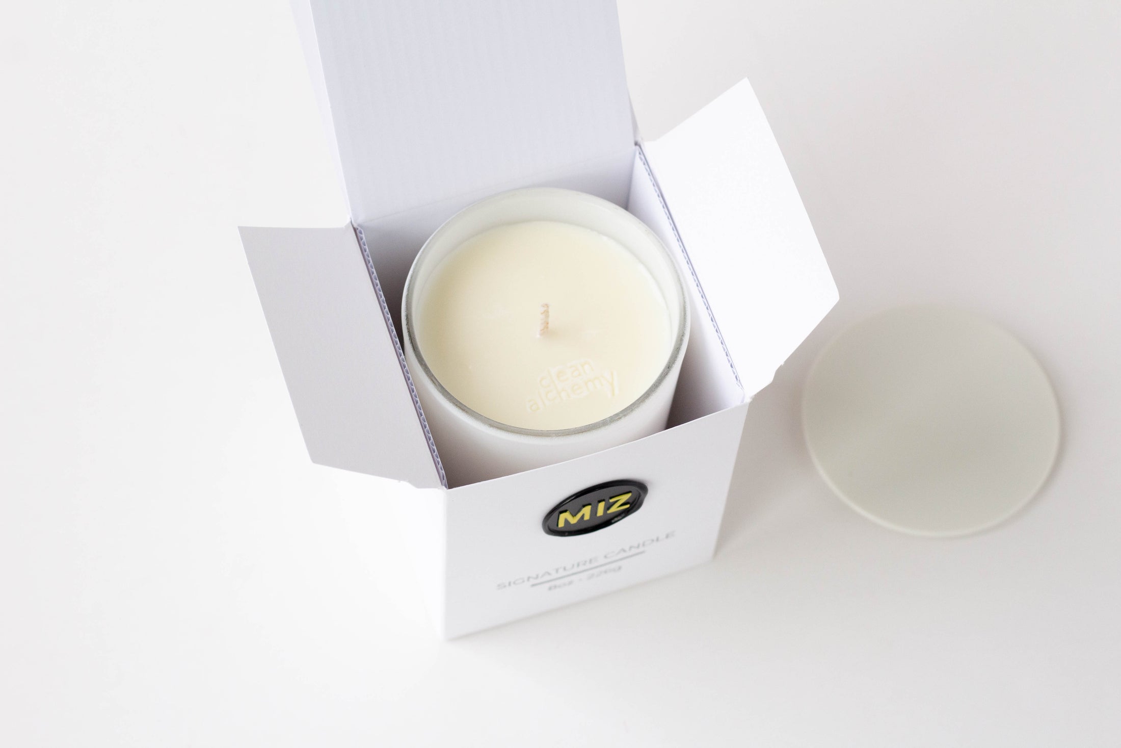 An open white candle box containing a white wax candle, with the brand logo 'MIZ' visible on the front. A matching white lid sits to the right on a clean white background.