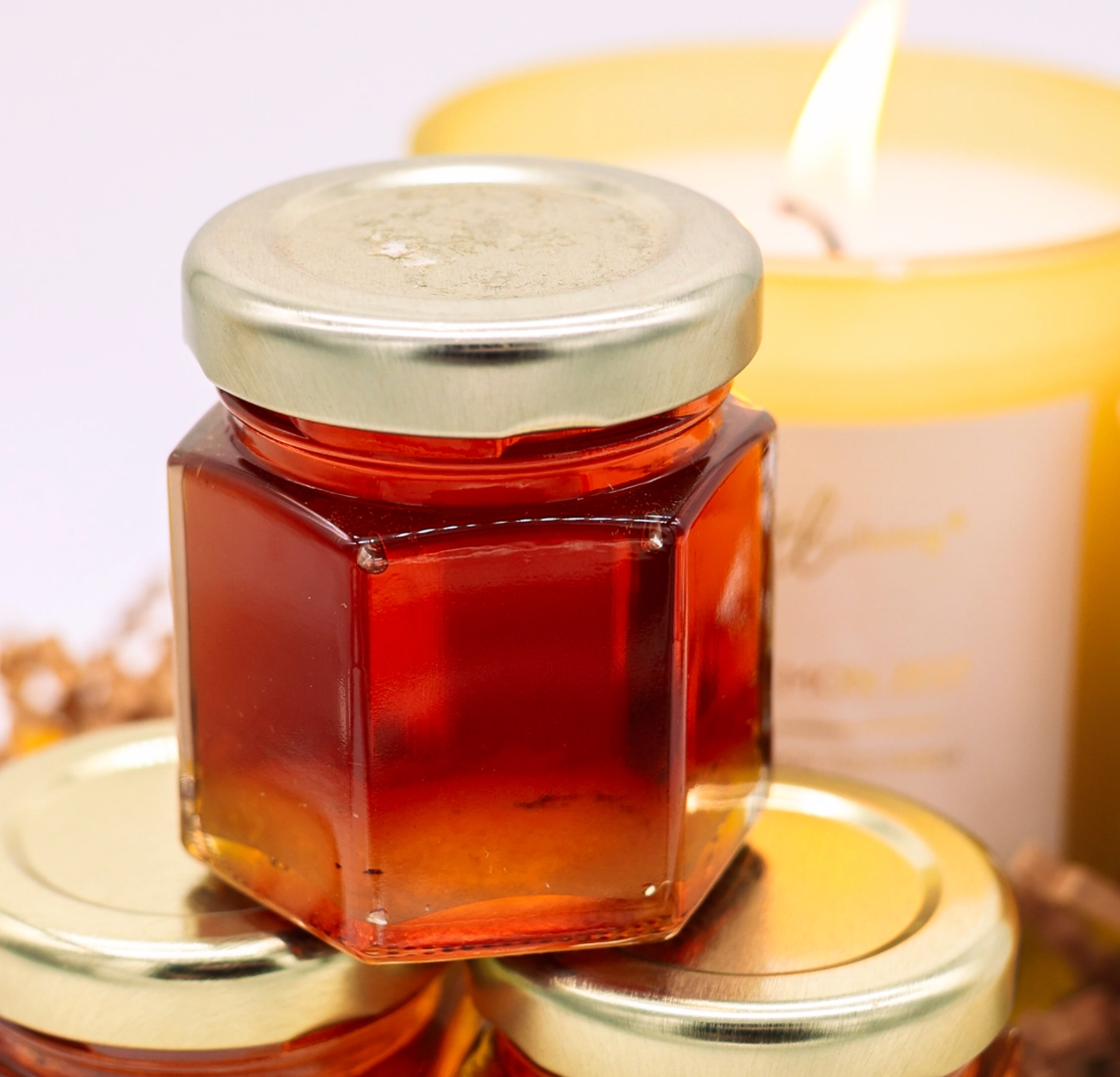 A hexagonal glass jar filled with amber-red honey, topped with a silver metal lid, displayed on top of other similar jars with a softly blurred yellow candle in the background.