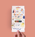 Load image into Gallery viewer, A hand holds the packaging for scrollino coloring book, filled with crayons and 1.5yds of paper to color on. 
