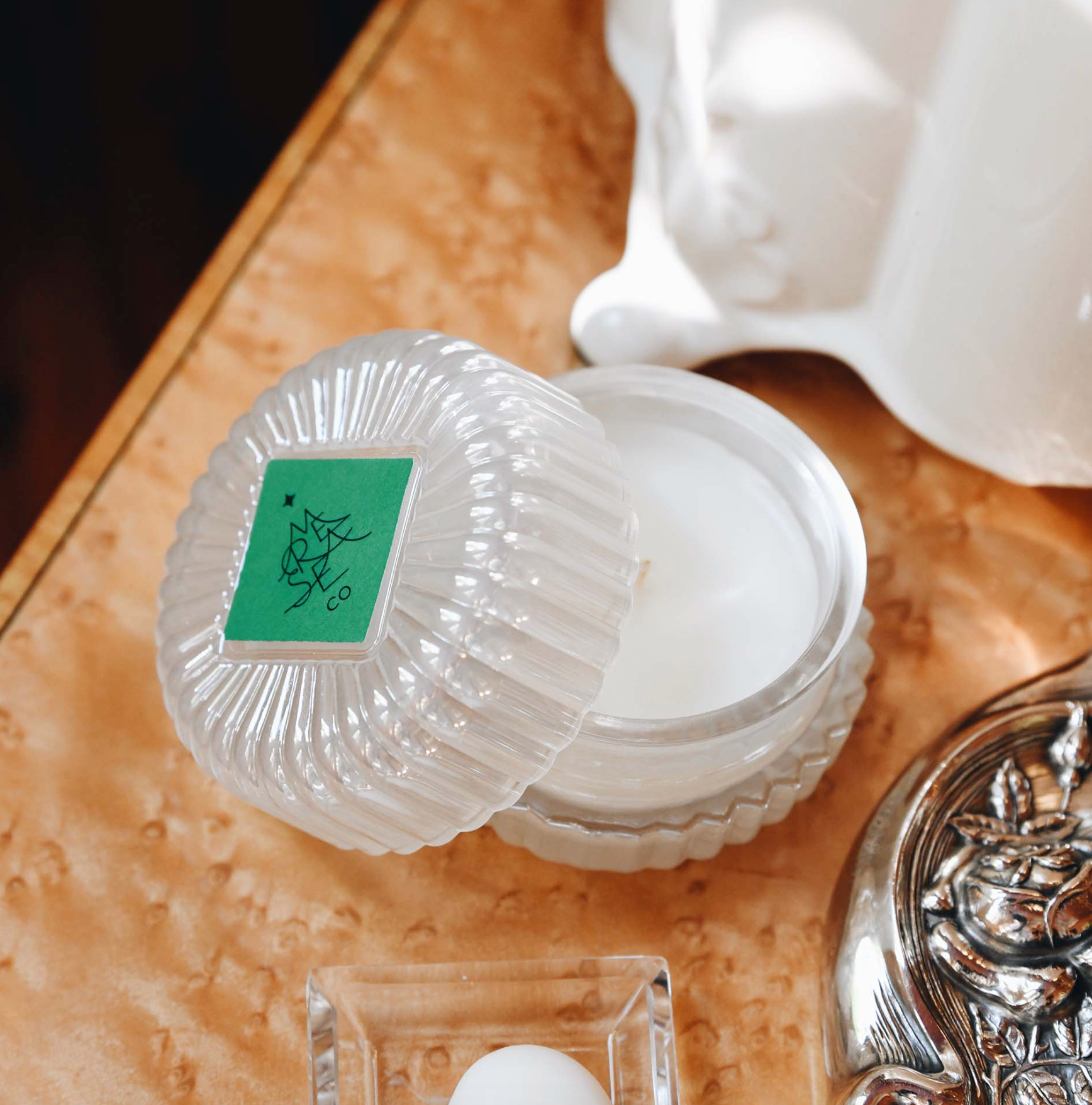 A clear ribbed glass jar candle with its lid off, showing white wax inside. The lid has a green square label with a black botanical-style logo.