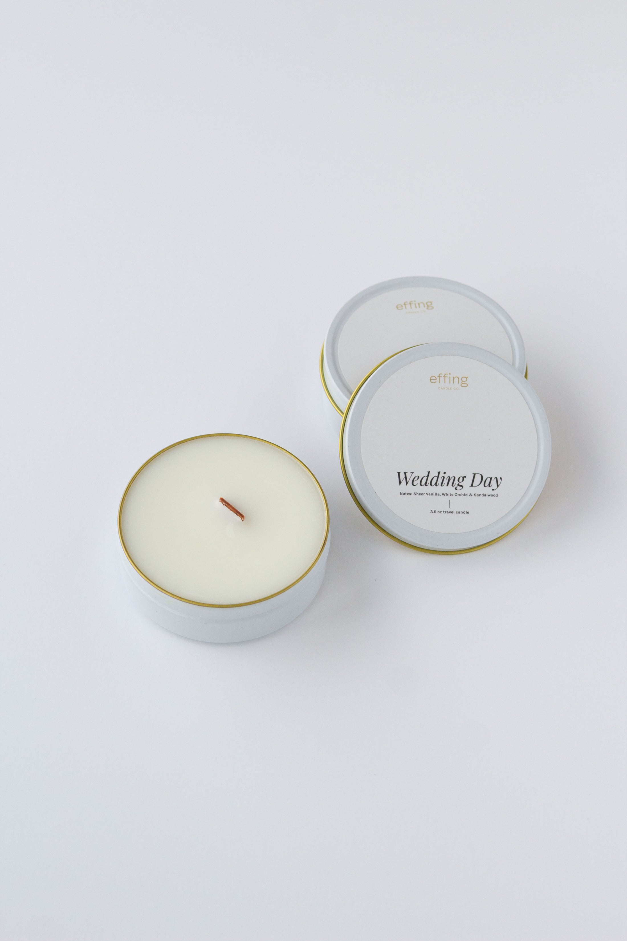 Two round candles in white metal tins with gold rims on a clean white background. One tin is open, showing pale cream wax and a single wood wick, while the other one is closed with white labels reading 'effing candle co' and 'Wedding Day'.