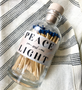Load image into Gallery viewer, A clear glass bottle filled with wooden matchsticks, sealed with a light wood cork, and labeled 'PEACE and LIGHT' in black text. The bottle is placed on a cream fabric with black woven stripes.
